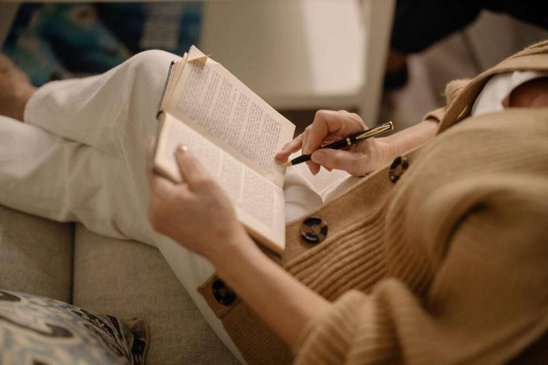 A woman seated on a couch, reading a book with a pen in her hand.