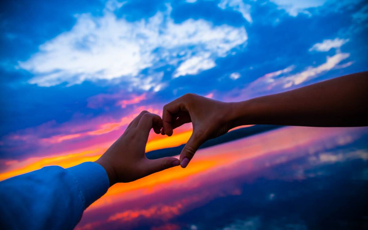 Two hands making a heart formation in front of a colorful, sunset horizon.