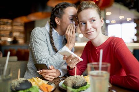 Two casual friendly girls with smartphones gossipping by served table in cafe.
