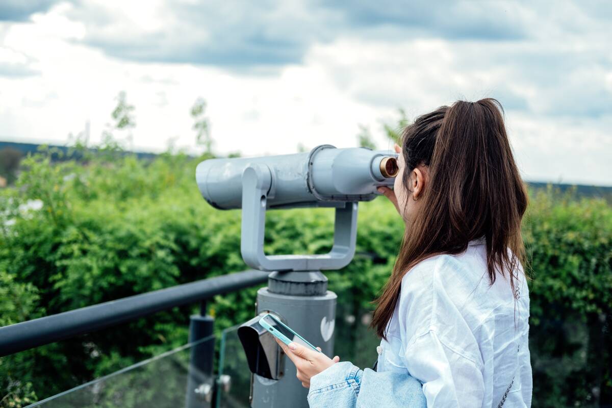Young woman tourist looking in binoculars, telescope in the city view. Back view of woman use binoscope in the viewpoint on the river bank.