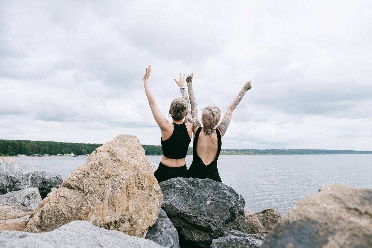 Two friends sitting on a large rock on the shore, raising their arms in celebration.