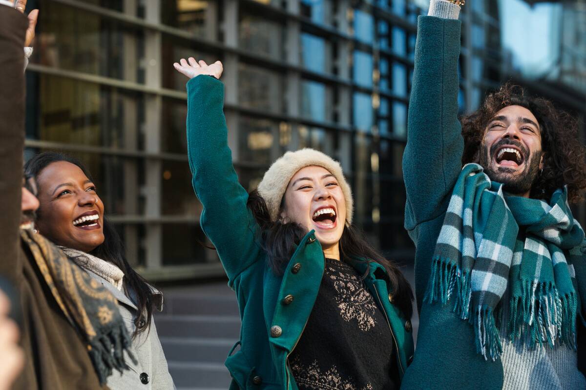 Cheerful man and women in outerwear raising arms and laughing after stacking hands while celebrating success on street together.