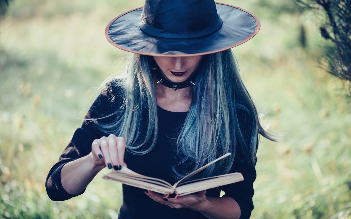 A woman with long hair, a witch hat, and black clothing smirking as she looks down into a book she's holding.