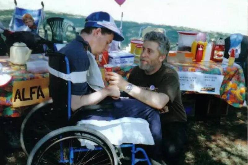 martin sitting in his wheelchair while out at an event, his dad helping him drink.