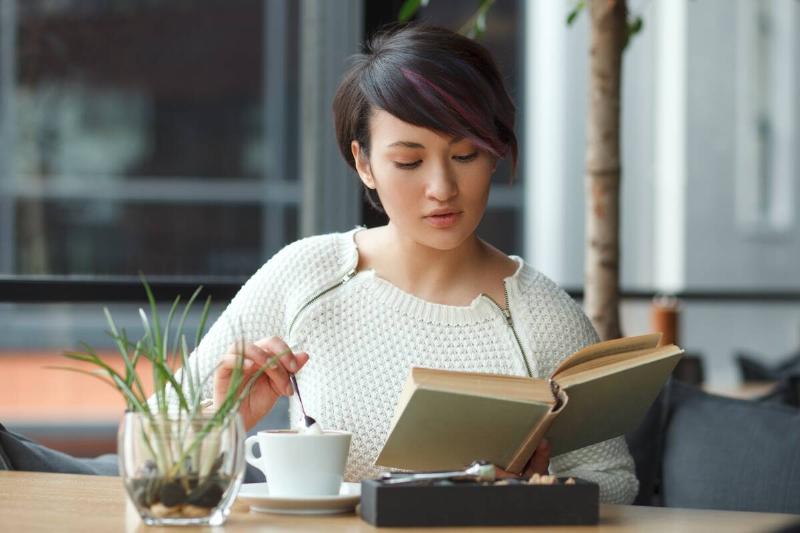 Young elegant woman sitting at table stirring up coffee with spoon and reading book in cafe.