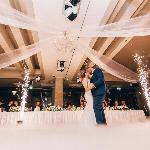 A low-angle shot of a couple kissing at a wedding, sparklers flying around them and a set table full of friends behind them.