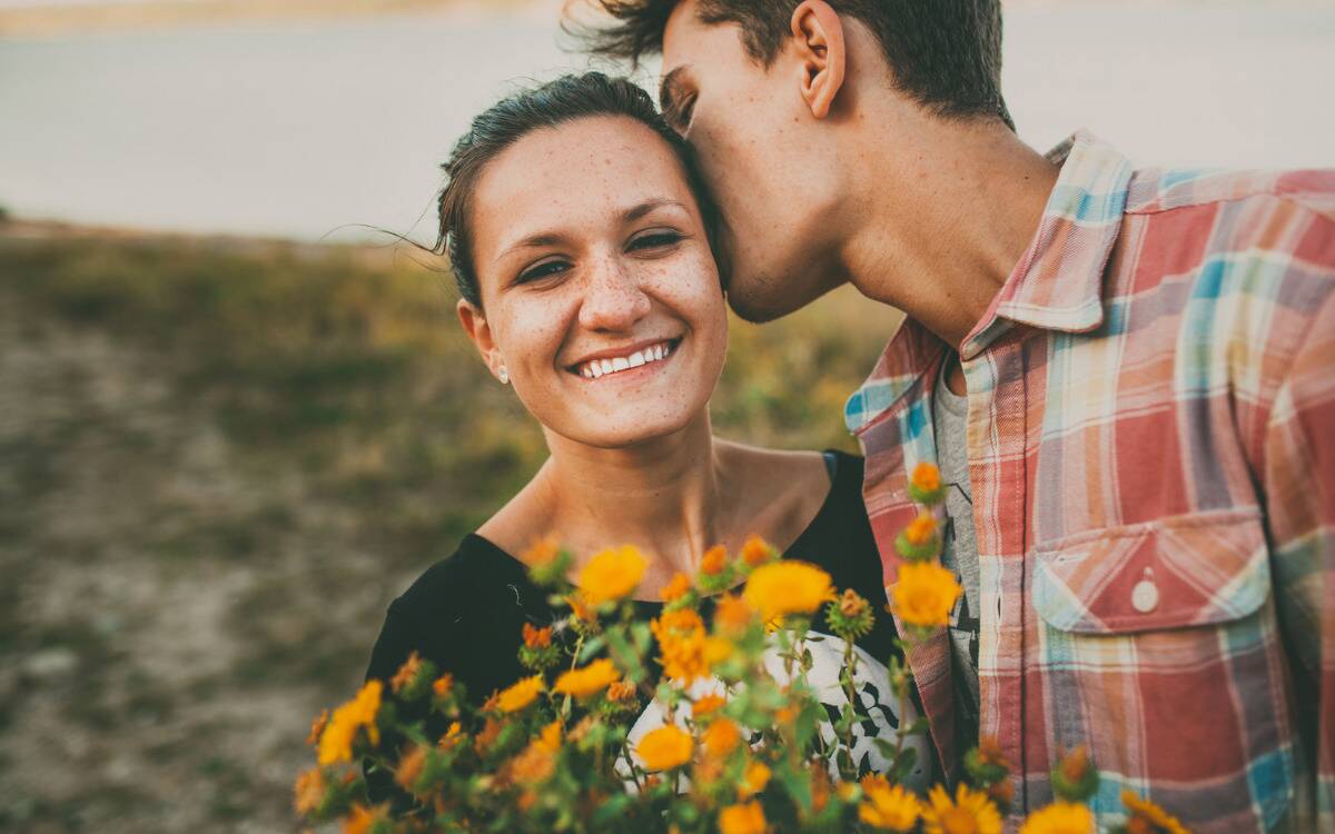 A woman smiling as her boyfriend kisses her head, a bouquet of yellow wildflowers in front of them.