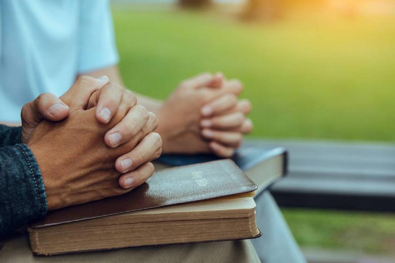 people seated outdoors legs crossed with hands folded over bible on lap