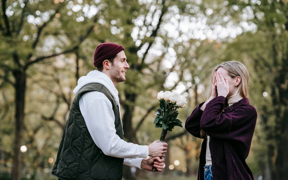 A couple standing in a park, the woman covering her eyes with her hands while the man hands her a bouquet.