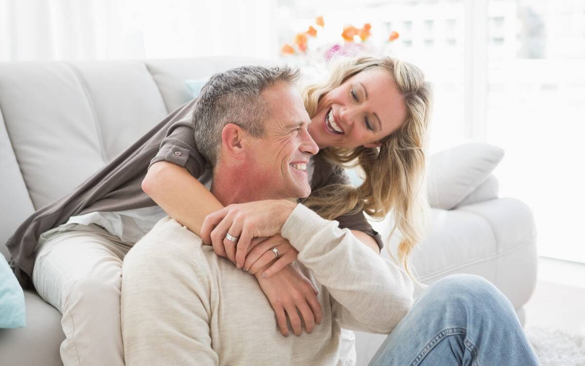 A woman sitting on a couch hugging her husband from behind, who's sitting on the ground in front of the couch.