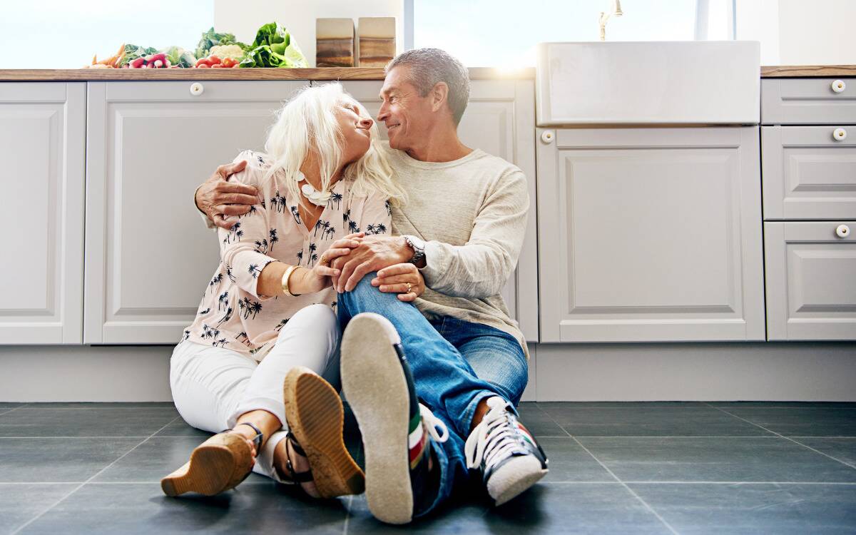 A couple sitting in each other's embrace on their kitchen floor, leaning against the lower cabinets.