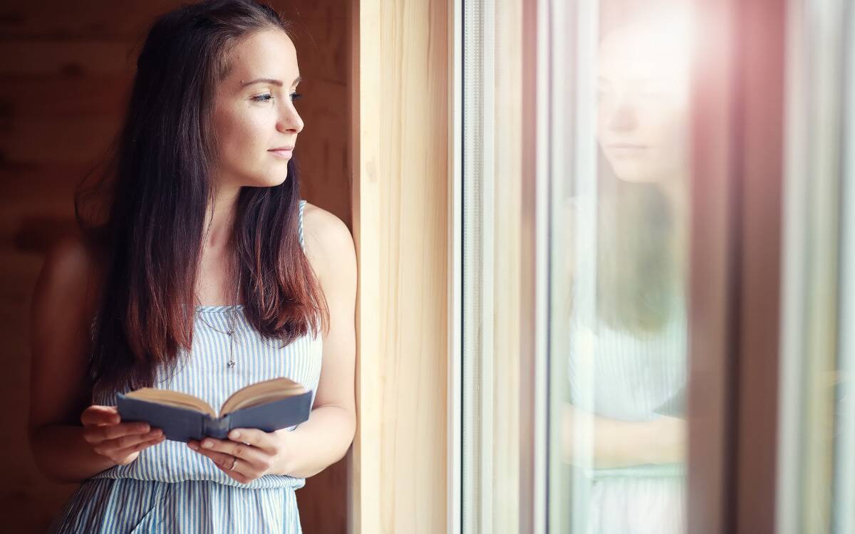 A woman holding a book as she looks out a window she's standing next to.