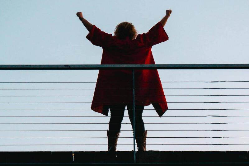 A woman in a red jacket standing on a bridge, arms up in celebration.