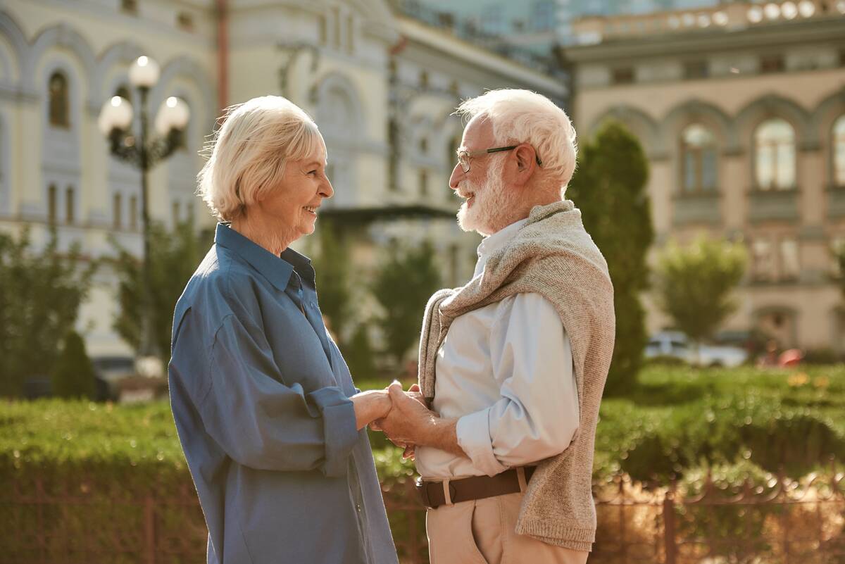 Elderly couple holding hands and looking at each other with smiles while standing outdoors.