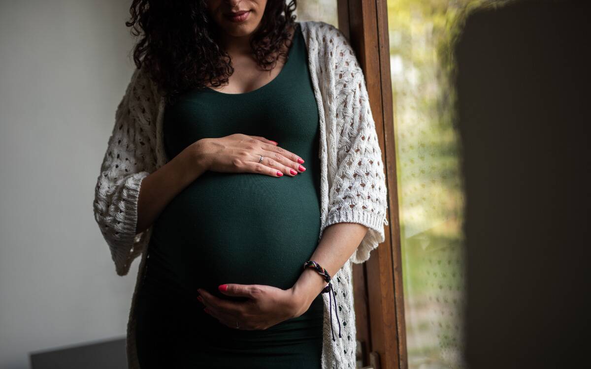 A pregnant woman in a green dress caressing her stomach next to a window.