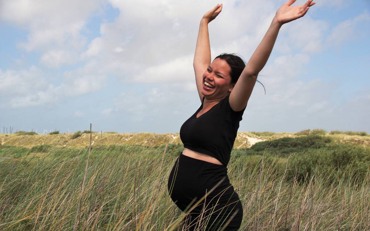 A pregnant woman standing in a field, arms raised above her head as she cheers.