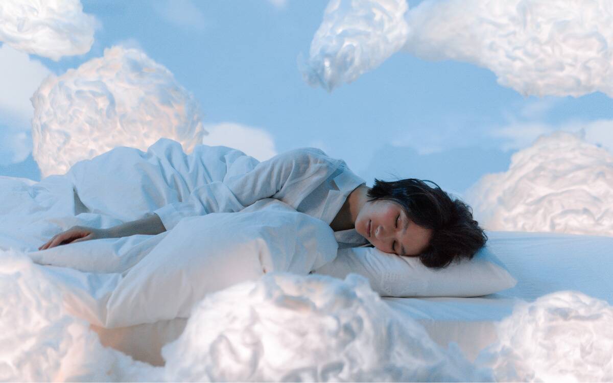 A woman laying down on a bed, wrapped in a blanket, surrounded by blue sky and clouds.