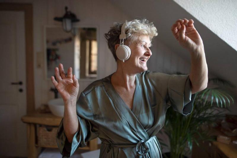 An older woman dancing by herself in her room, wearing headphones and a silk robe.