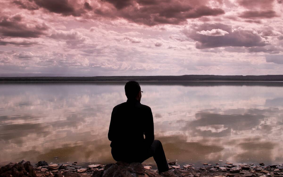 A silhouette of a man sitting on a rock in front of the water, the sky a pink/purple hue.