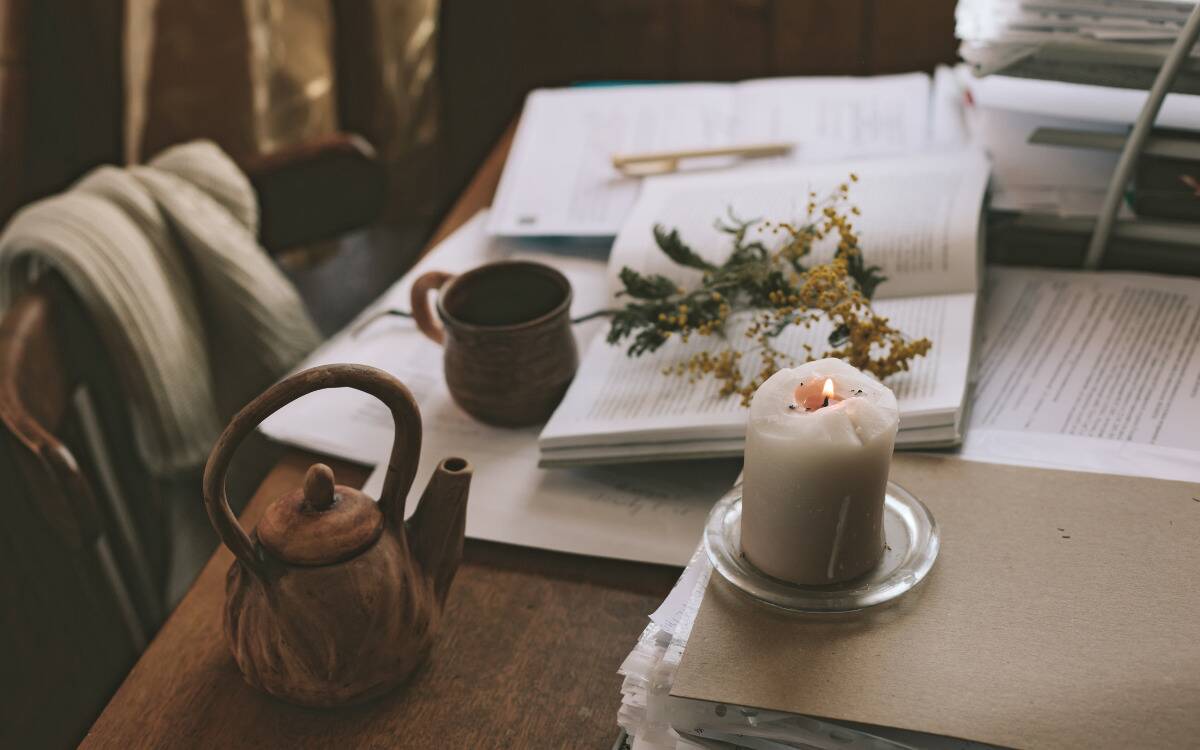 A home table covered in books, papers, flowers, a candle, a teapot, and a mug.