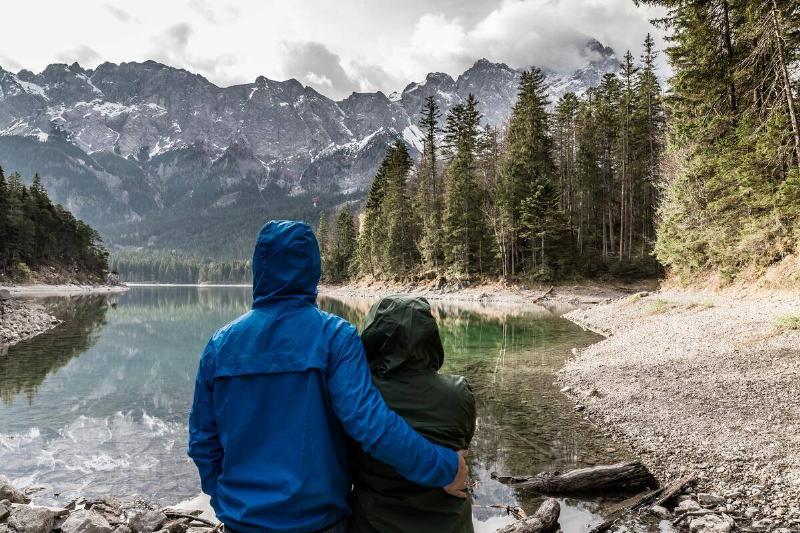 Two people in raincoats standing in front of a lake at the base of a mountain range.
