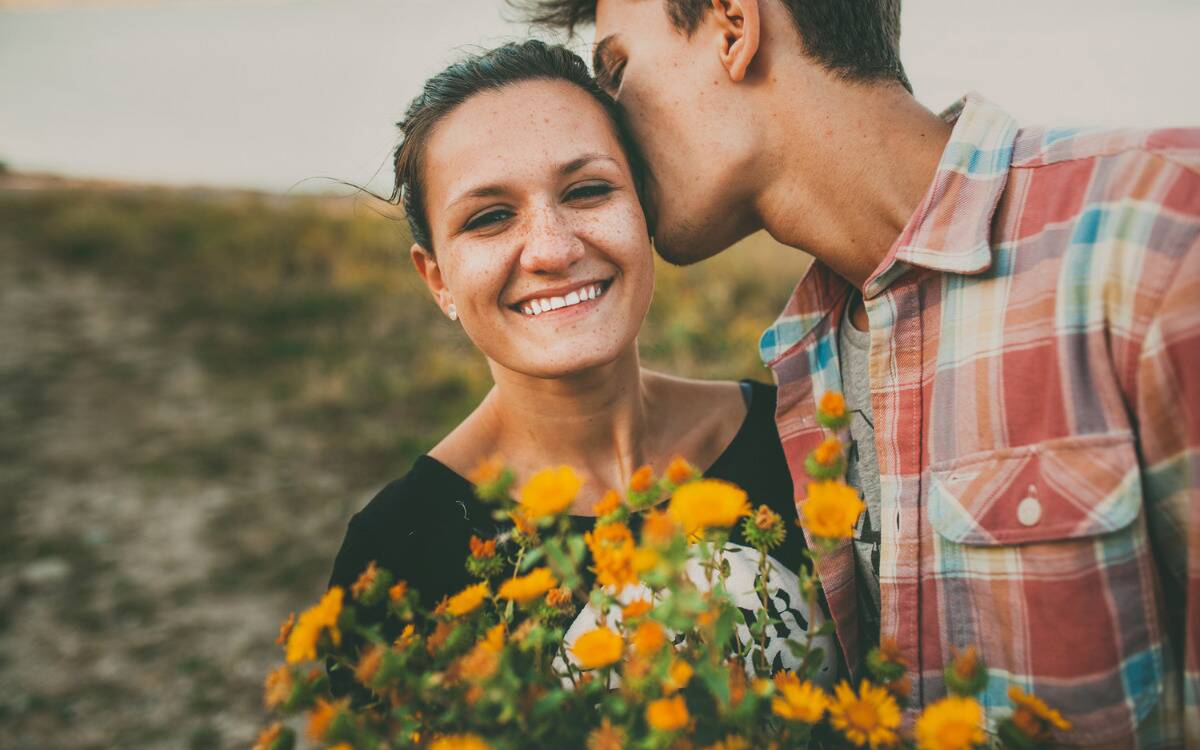 A couple standign in a field, the woman holding a bouquet while the man kisses the side of her head.
