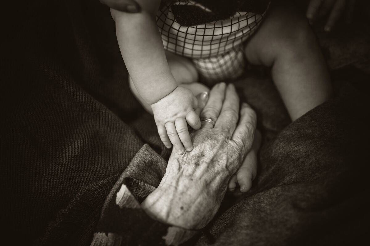 A greyscale image of an elderly person's hand resting near a baby's arm.