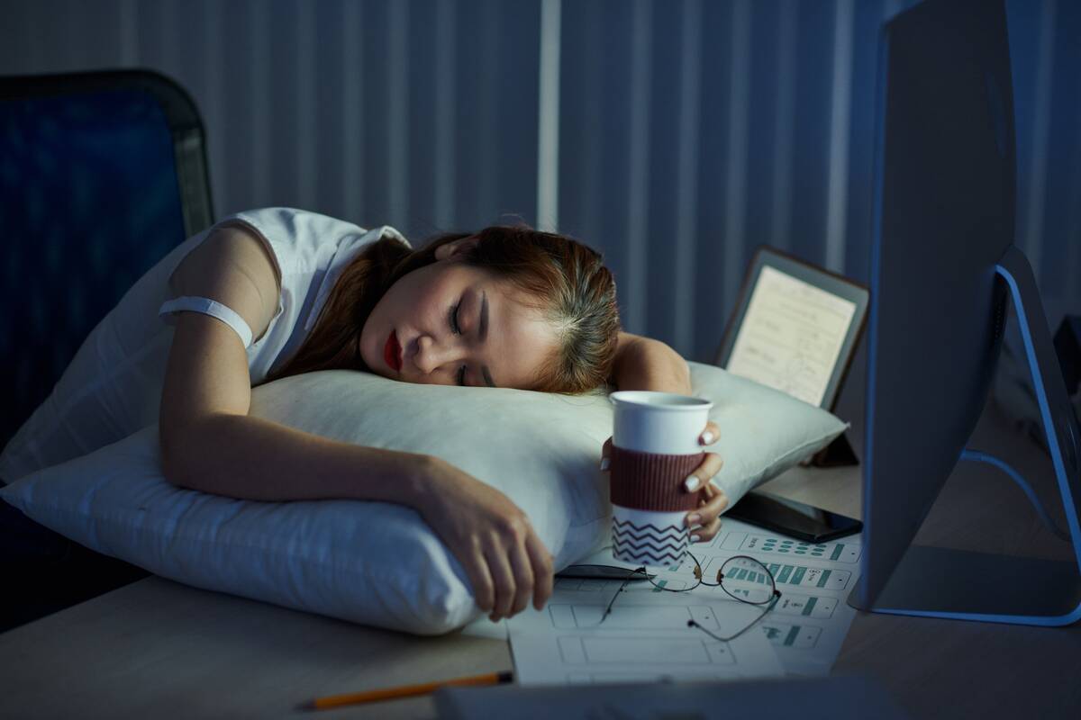 Tired young female employee fell asleep at office desk with coffee cup in her hands.