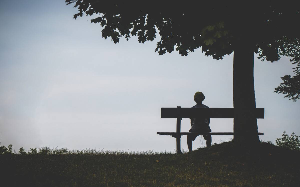 A silhouette of someone sitting on a bench alone under a tree.