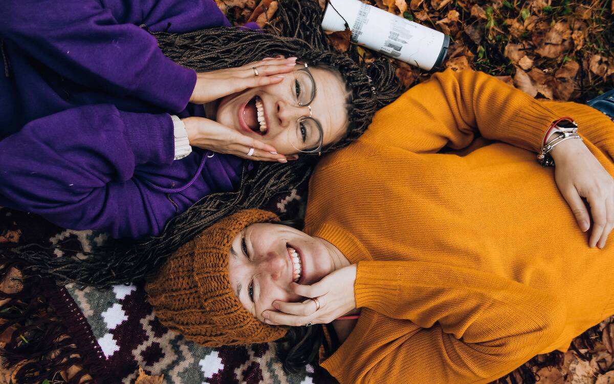 Two friends laying in the autumn leaves, their heads next to each other, smiling up at the camera.