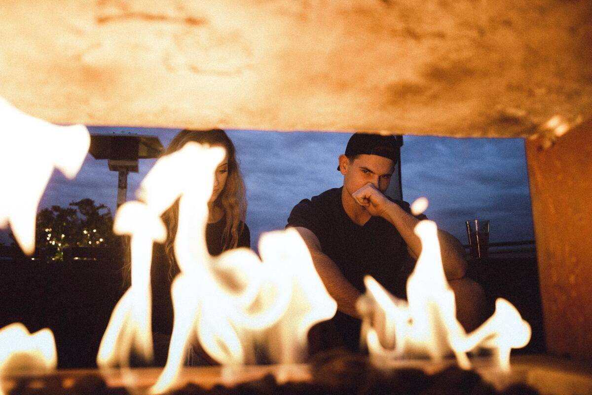 A couple sitting otuside, looking tense, seen through the flames of a fire.