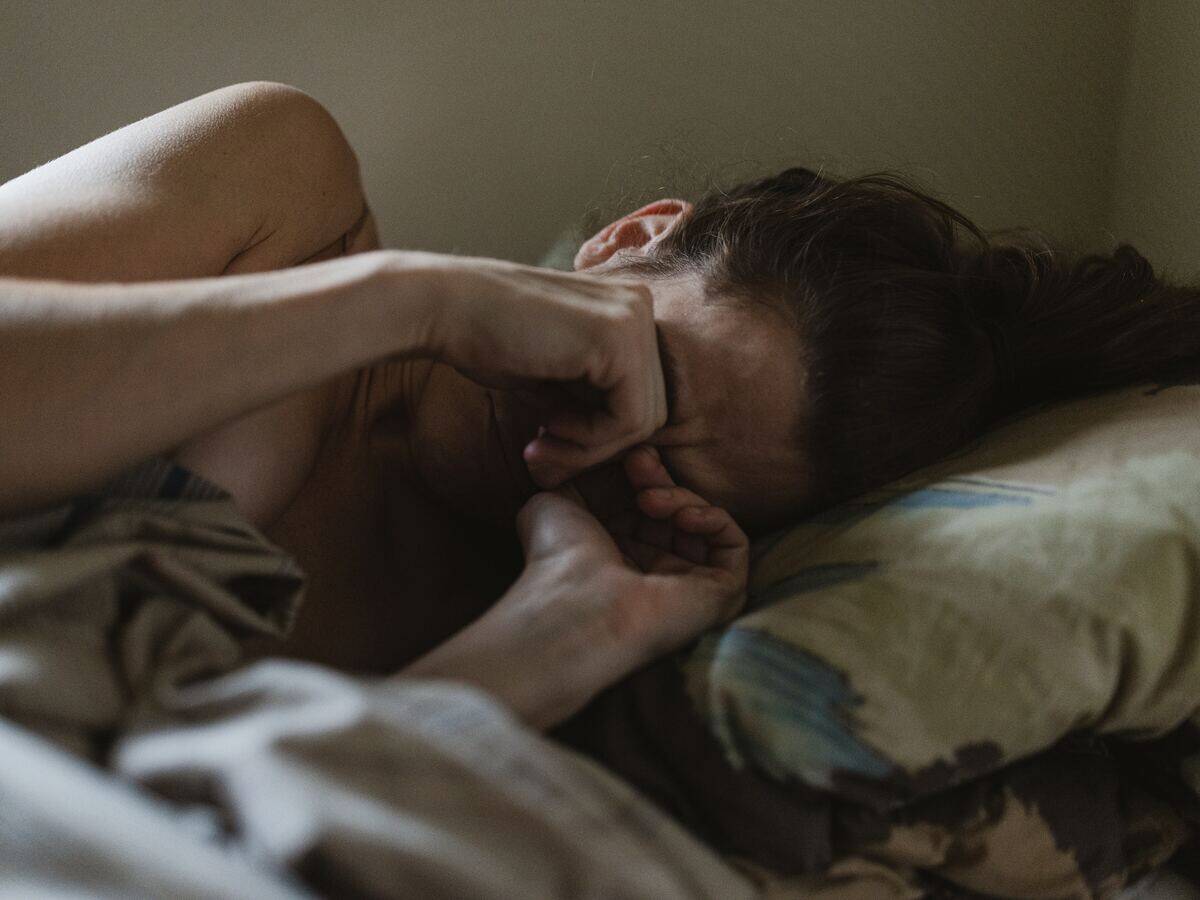 A woman laying in bed, face scrunched up, hands covering her eyes as she cries.