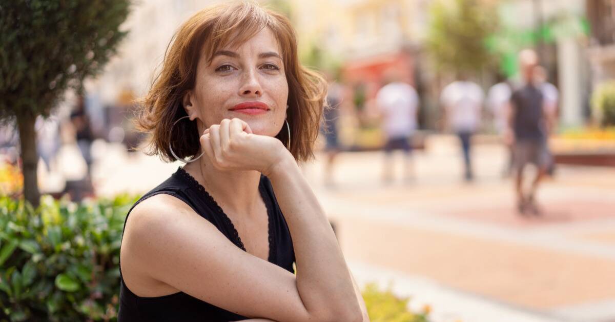 An elegant woman sitting outside, looking at the camera, resting her chin on her hand.