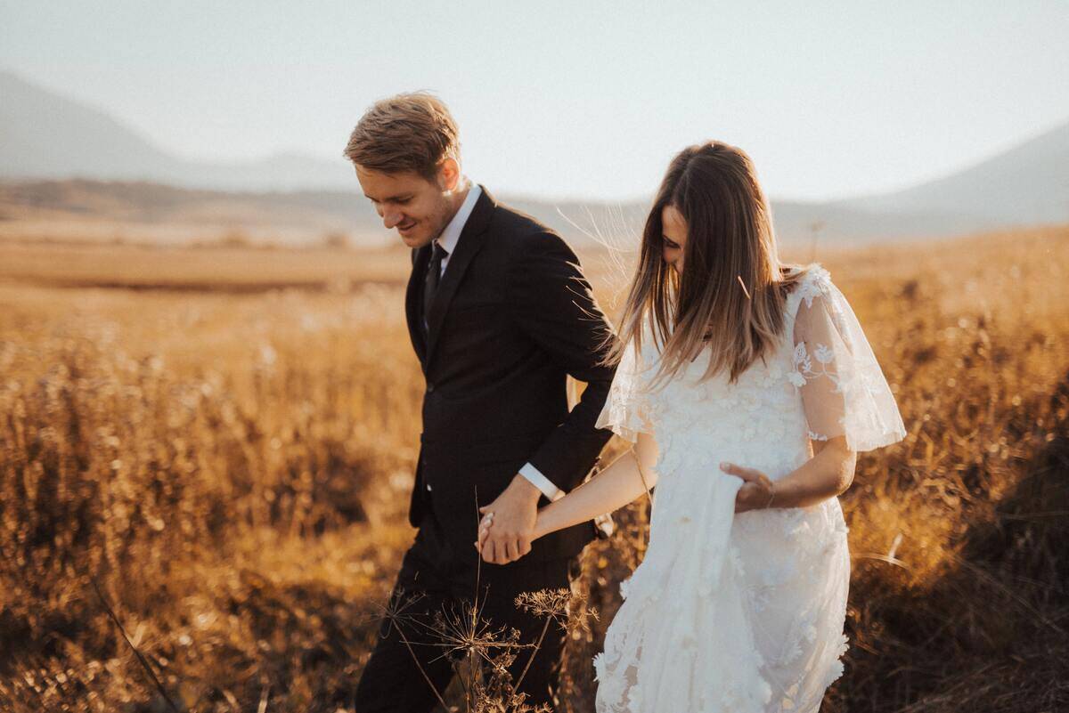A young married couple walking through a field, holding hands, still in their clothing from the wedding.