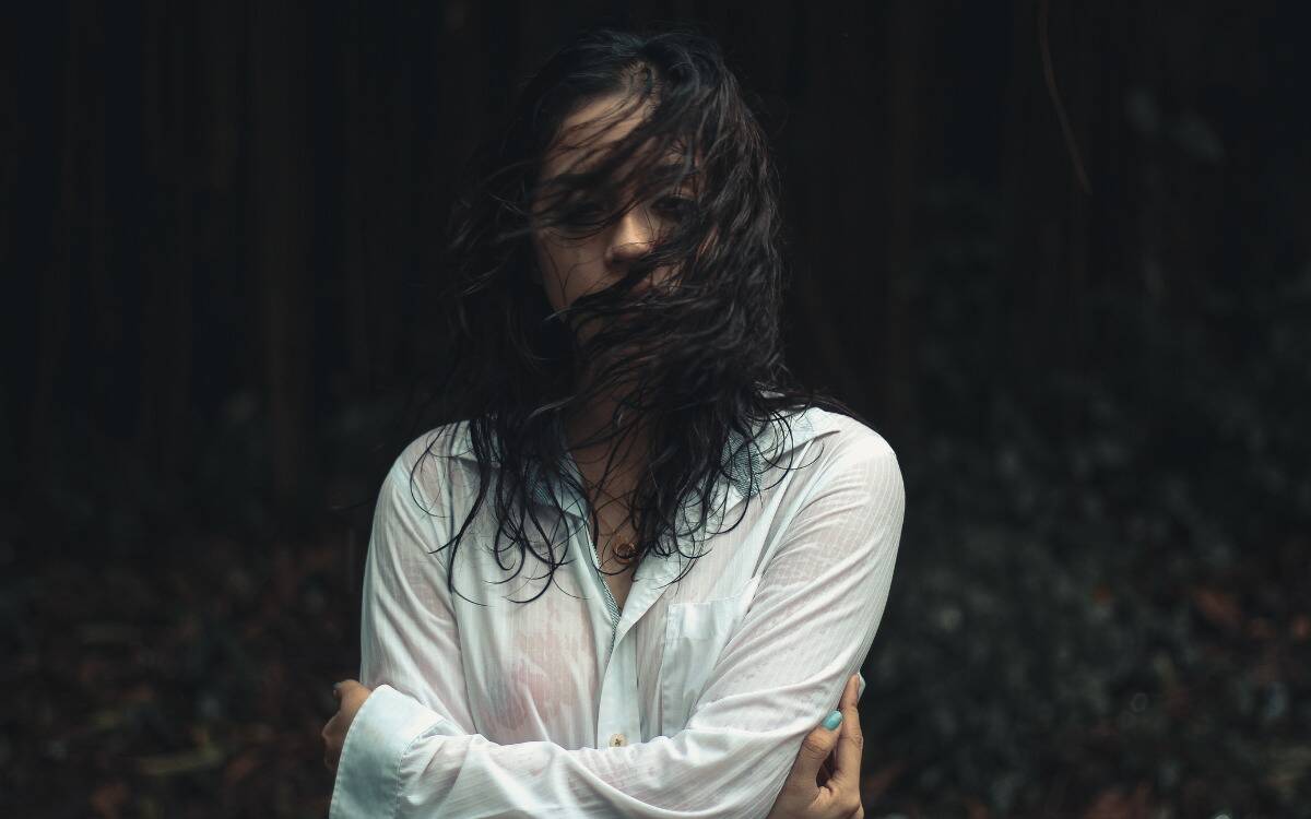 A woman standing in a dark scene, her hair blowing in front of her face, appearing wet from the rain.