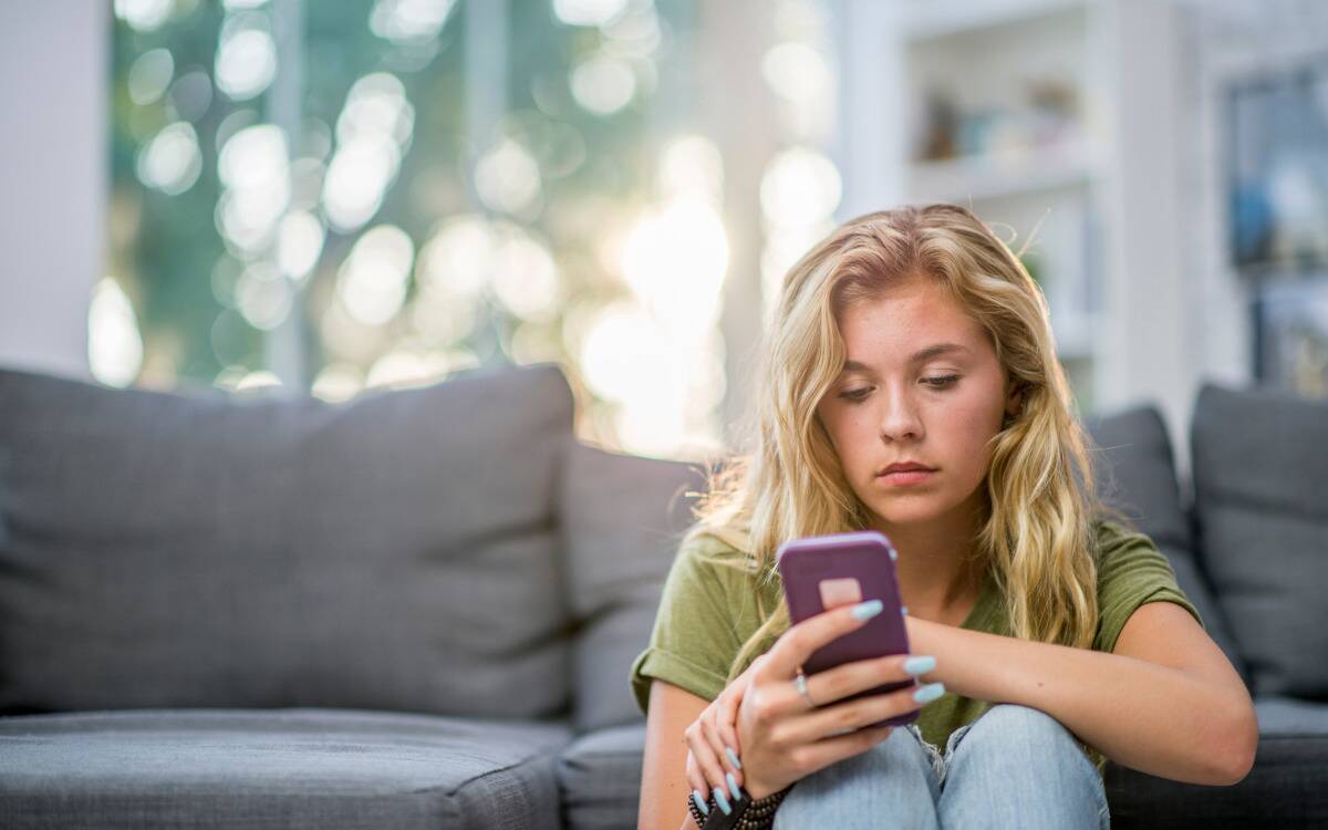 A woman sitting in front of her couch looking at her phone dejectedly. 