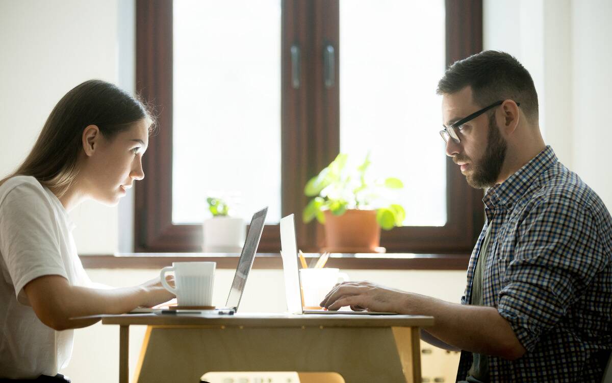 A couple sitting at a table, each at a laptop, looking at each other incredulously. 