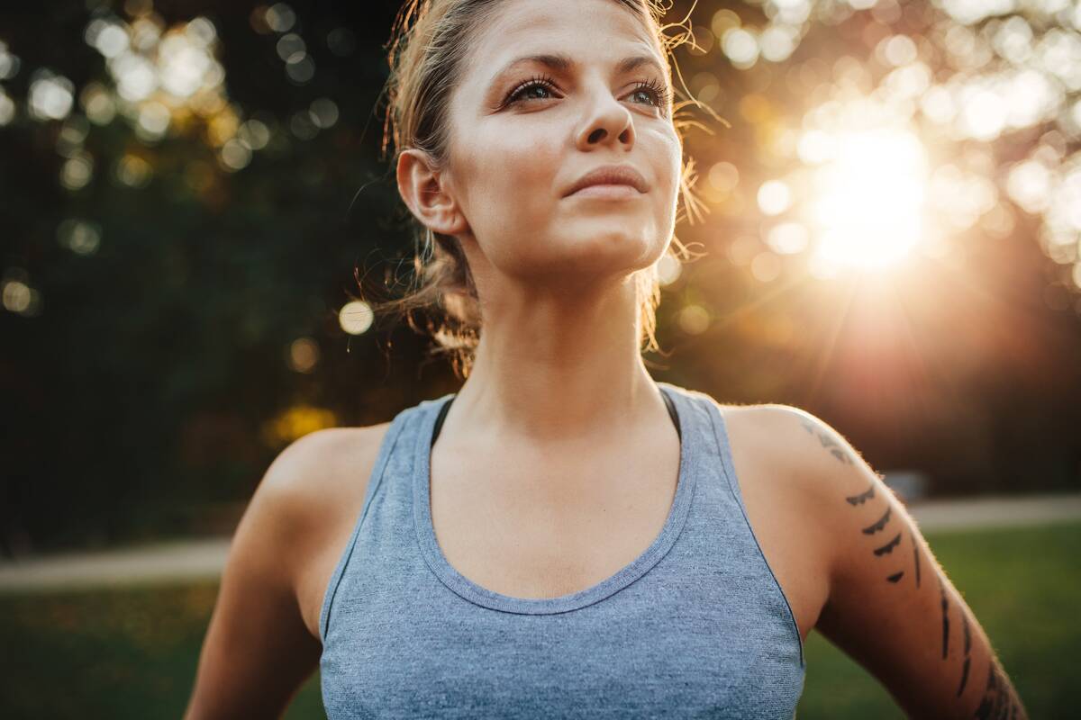 A closeup shot of a woman standing in a park, looking confident, the sun shining behind her.