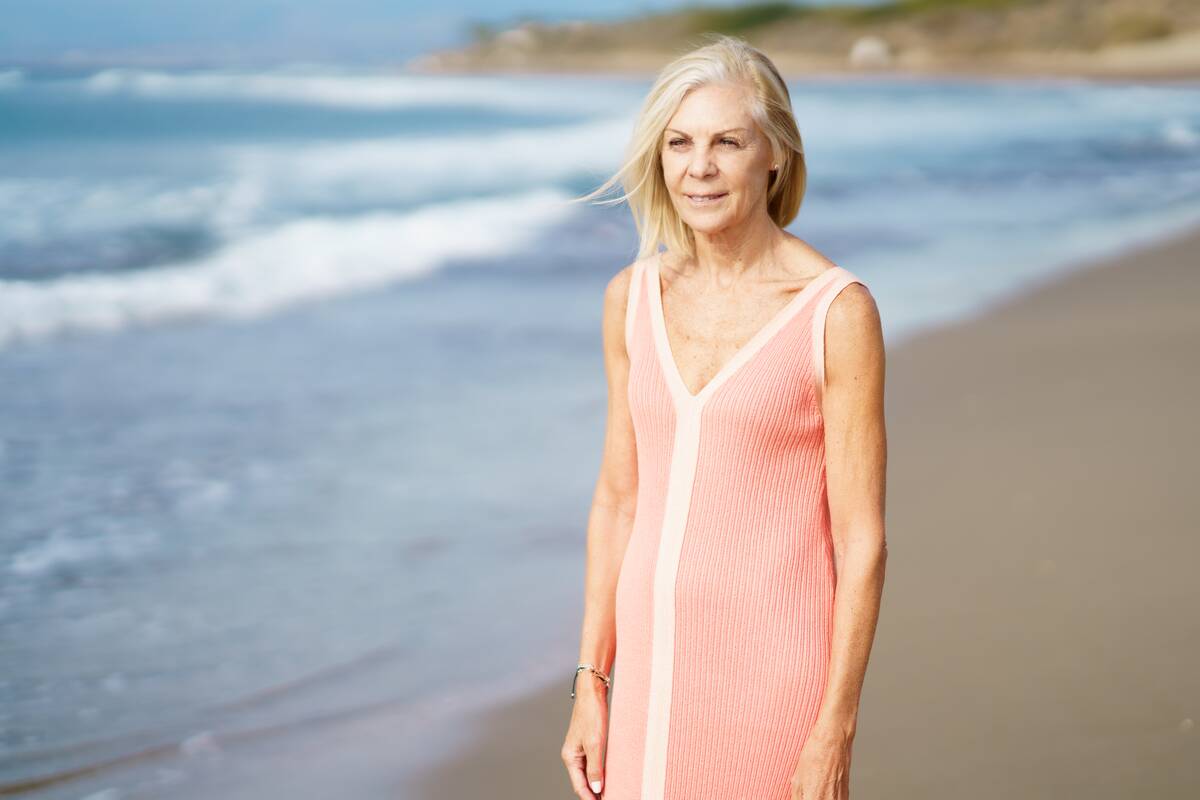 Serene senior female in dress and with gray hair standing on sandy coast near sea in summer and looking away.