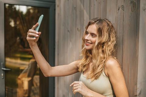 A young woman taking a selfie as she leans against a wooden wall.