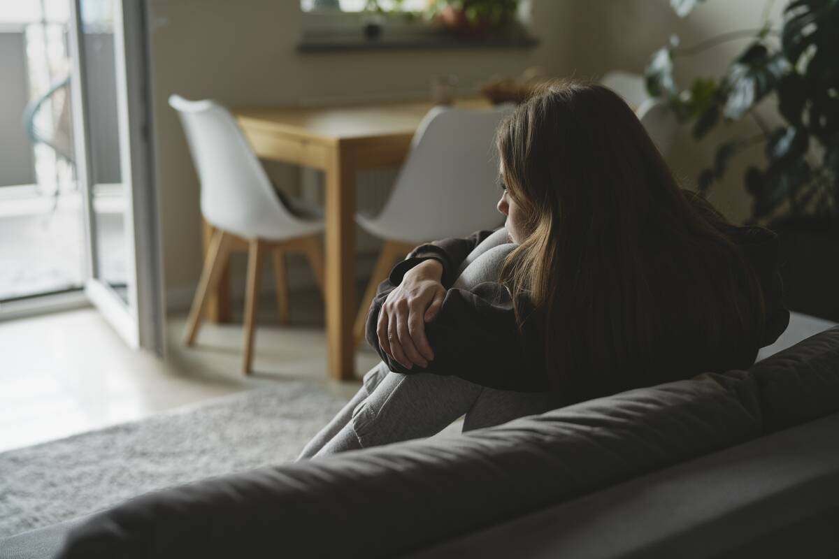 A woman sitting on her couch, knees drawn to her chest, looking sad.