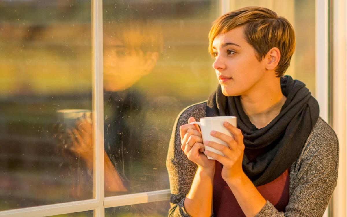 A woman sitting with a mug in her hands, looking longingly out a window.