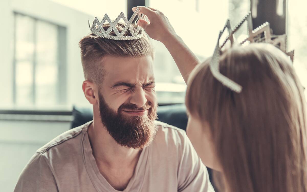 A father scrunching up his face as his daughter places a tiara on his head.