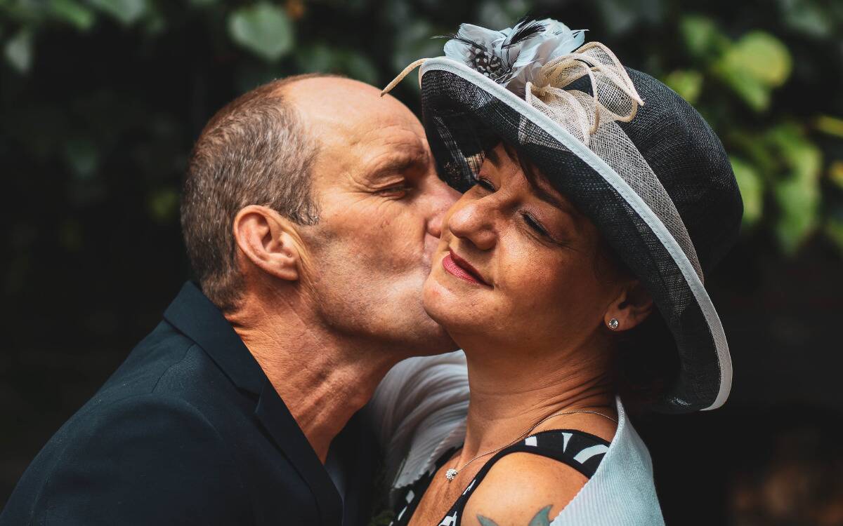 A couple standing close to one another, the man kissing the woman on the cheek, the woman in a fancy hat.