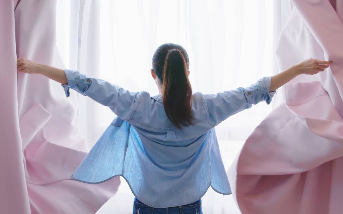 A woman faced away from the camera in front of a window, throwing the curtains open.