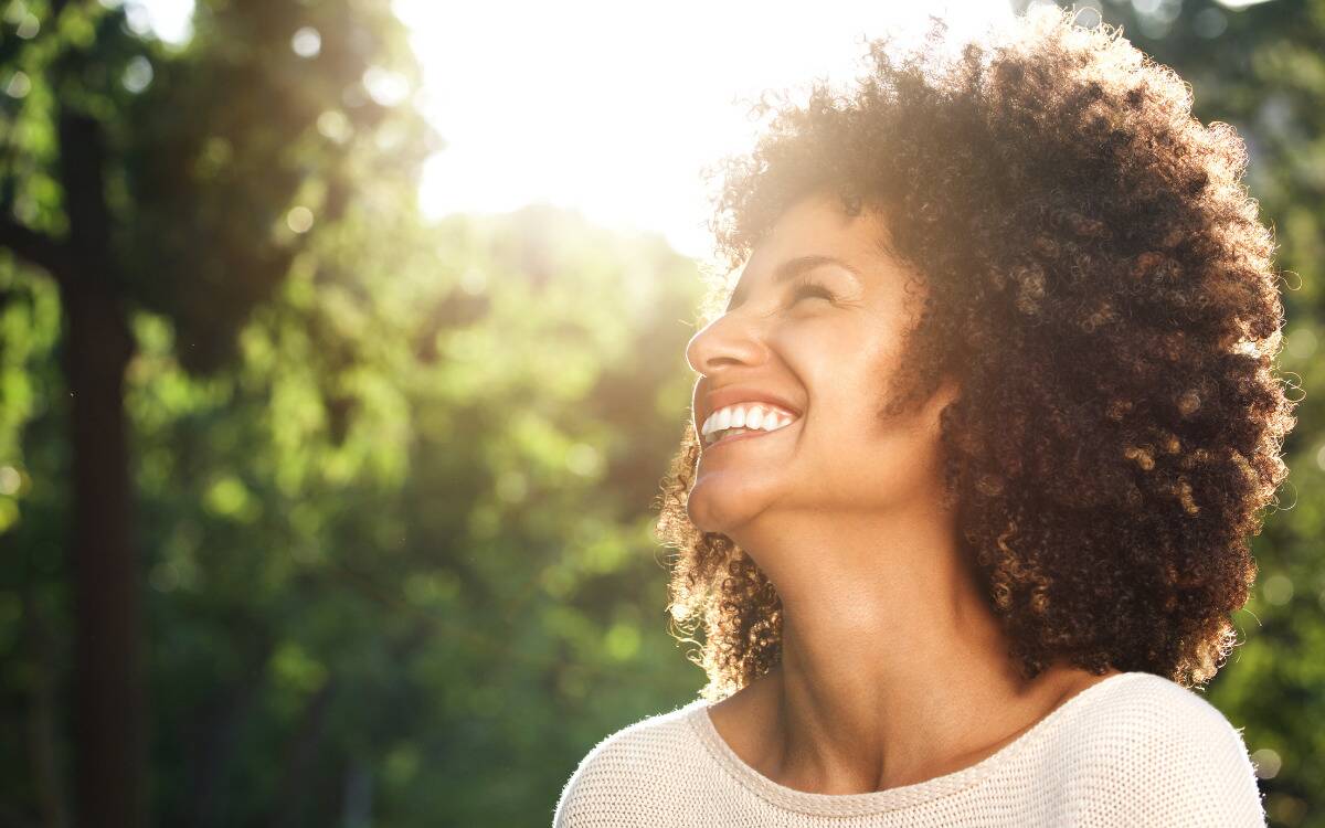 A woman smiling up at the sunshine as she stands outside.