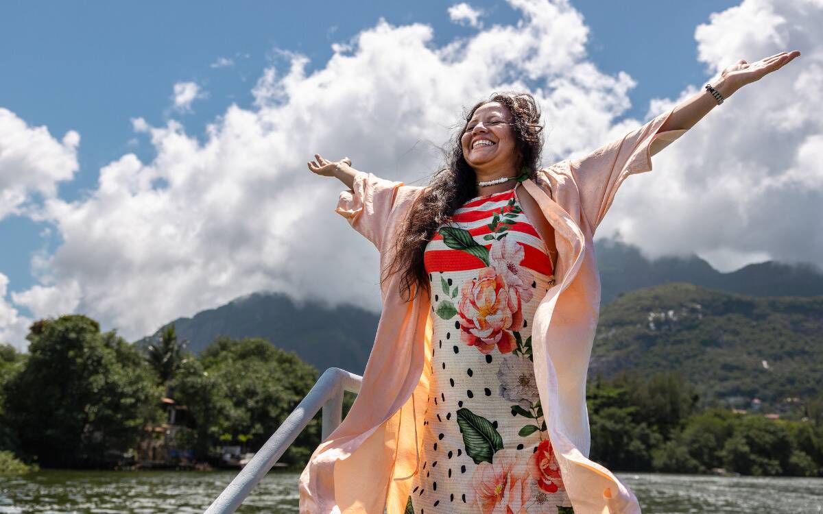 A woman in a colorful dress standing by the water with her arms closed, arms up, basking in the sunlight.