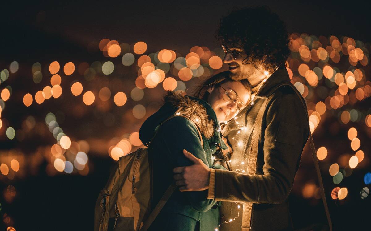 A couple embracing, the woman resting her head on the man's chest, both surrounded by fairy lights.