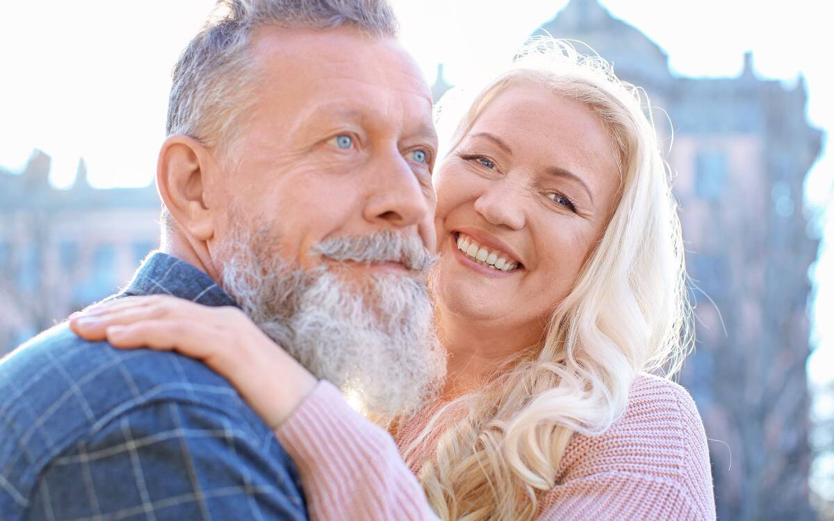 A mature couple embracing, the woman looking at the camera and the man looking off in the distance.