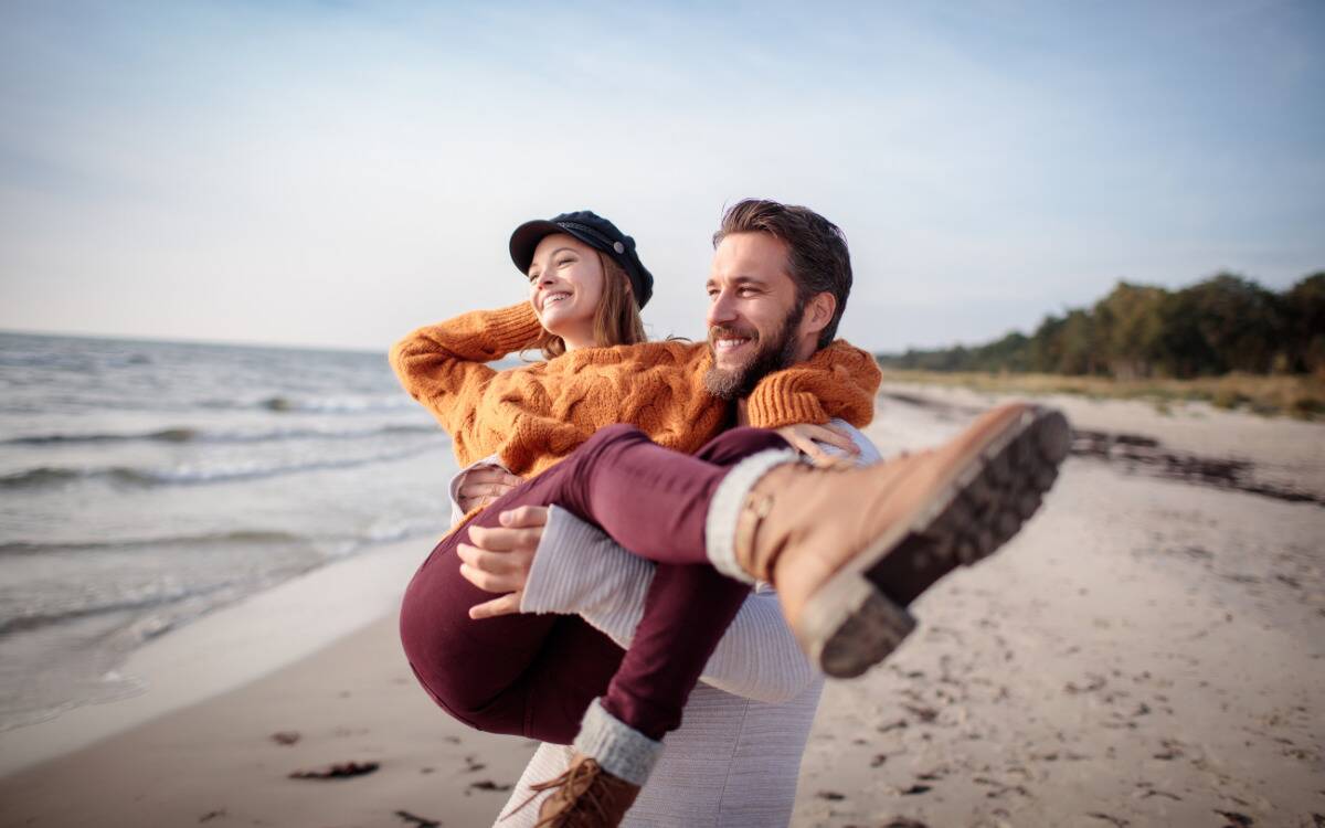 A couple on the beach, the man carrying the woman bridal style as they look out at the water.